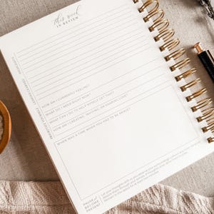May include: A white journal with a gold spiral binding. The cover has the text "this week in review" written in cursive. The journal has lined pages with prompts for reflection and self-care.