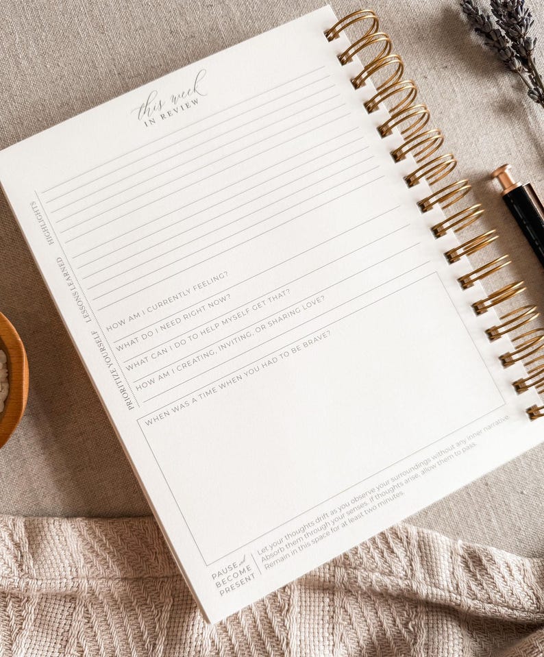 May include: A white journal with a gold spiral binding. The cover has the text "this week in review" written in cursive. The journal has lined pages with prompts for reflection and self-care.