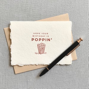 May include: A white card with a brown border and the text "HOPE YOUR BIRTHDAY IS POPPIN'" in brown lettering. A red illustration of a popcorn bucket with popcorn inside is below the text. A black pen with a gold top is lying on the card.