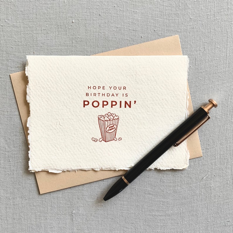 May include: A white card with a brown border and the text "HOPE YOUR BIRTHDAY IS POPPIN'" in brown lettering. A red illustration of a popcorn bucket with popcorn inside is below the text. A black pen with a gold top is lying on the card.