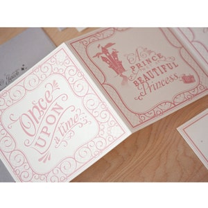 May include: A pink and white folded card with a pink ornate border. The front of the card says "Once Upon a Time..." and the back says "A Prince met a Beautiful Princess".