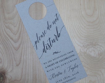 May include: A brown paper door hanger with a hole at the top. The text "please do not disturb" is written in black cursive script. Below the text is a small sprig of leaves. The text "We are recovering from a night of celebrating with the newlyweds Kristin & Taylor June 18th, 2016 #KTWEDDING" is written in black text.