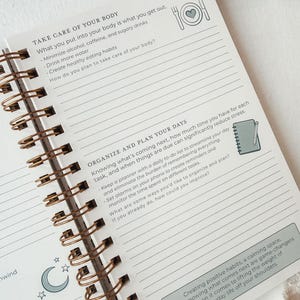 Pu&ograve; includere: Una pagina di agenda con rilegatura a spirale che presenta le sezioni TAKE CARE OF YOUR BODY e ORGANIZE AND PLAN YOUR DAYS. La pagina include aree di scrittura a righe, icone di un piatto con un cuore, un taccuino con una matita e una luna.
