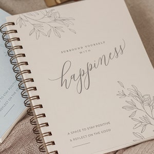 May include: A white spiral bound journal with the text "Surround yourself with happiness" and a floral design. The journal has the text "A space to stay positive & reflect on the good" at the bottom.