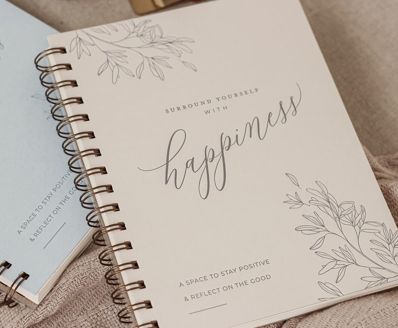 May include: A white spiral bound journal with the text "Surround yourself with happiness" and a floral design. The journal has the text "A space to stay positive & reflect on the good" at the bottom.