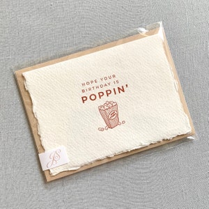 May include: A white card with a brown border and a red illustration of a popcorn bucket. The text on the card reads "Hope your birthday is poppin'"