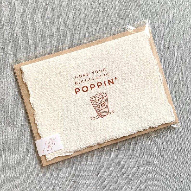 May include: A white card with a brown border and a red illustration of a popcorn bucket. The text on the card reads "Hope your birthday is poppin'"