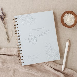 May include: A blue spiral bound journal with the text "Surround Yourself with happiness" and "A space to stay positive & reflect on the good". The journal is on a light brown surface with a small bowl of white salt and a white pen.
