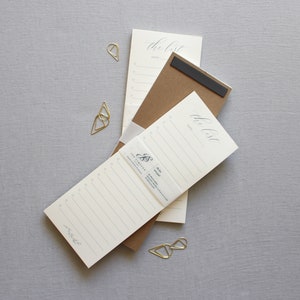 May include: Two white to-do list pads with the text "the list" and a date line. The pads are stacked on top of a brown pad with a black magnetic strip.