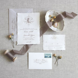 Op de afbeelding: Huwelijksuitnodigingsset met een witte achtergrond en bruine kalligrafie. De uitnodiging heeft een monogram met de initialen "K" en "L" in een decoratief lettertype. De RSVP-kaart heeft een decoratieve rand en de tekst "RSVP in the mail of love".