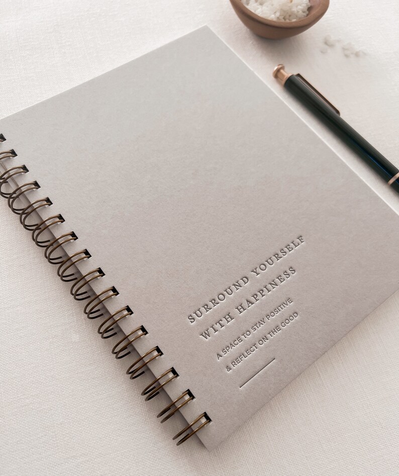 May include: A gray spiral bound journal with the text "Surround Yourself With Happiness" and "A space to stay positive & reflect on the good" printed on the cover.