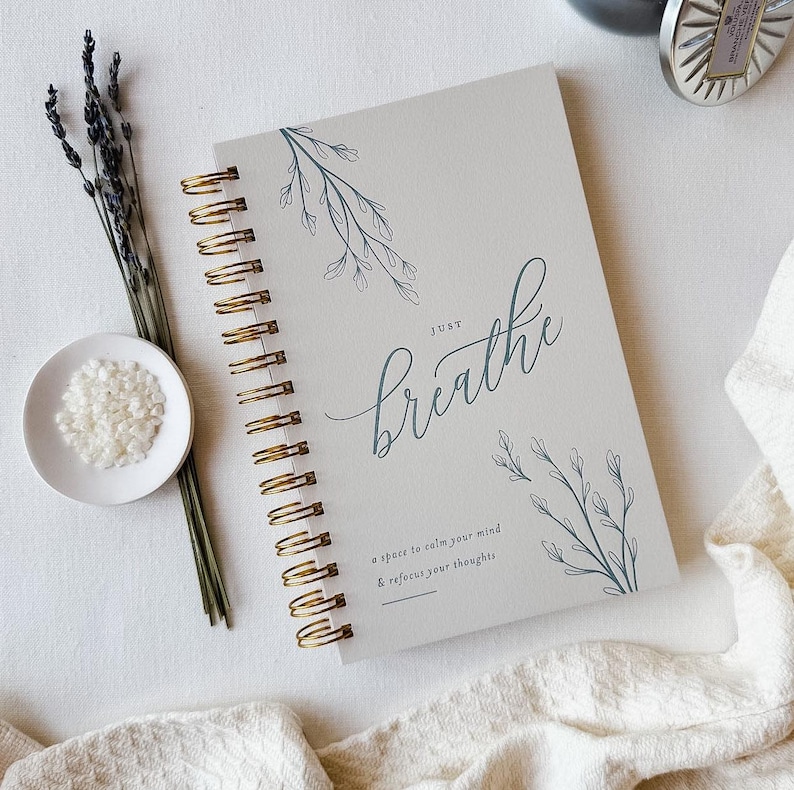 Pu&ograve; includere: Un taccuino a spirale con copertina grigio chiaro decorata con disegni botanici a linee color ottanio e il testo "JUST breathe" seguito da "a space to calm your mind & refocus your thoughts" in un carattere moderno con rilegatura dorata.