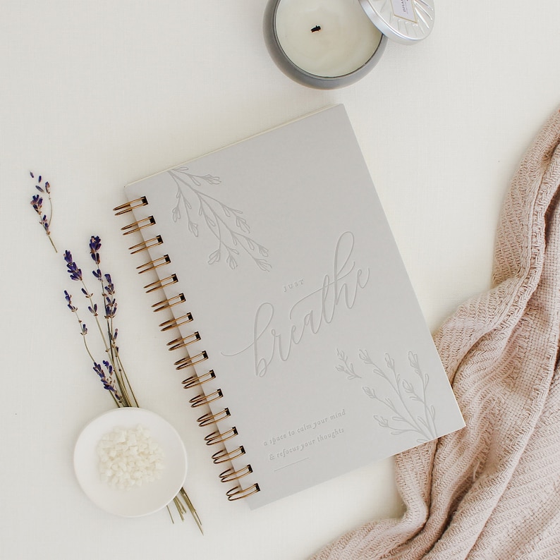 Pu&ograve; includere: Un taccuino a spirale grigio chiaro con rilegatura in filo metallico dorato. La copertina presenta disegni floreali in rilievo e il testo "JUST breathe" seguito da "a space to calm your mind & refocus your thoughts" in un carattere sans-serif.