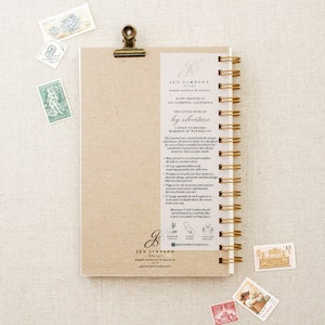 May include: A brown, spiral bound journal with a gold clip. The cover has a handwritten inscription that reads "The Little Book of Big Adventures: A Space to Record Memories of Wanderlust". The journal is designed to help you record your travel memories.