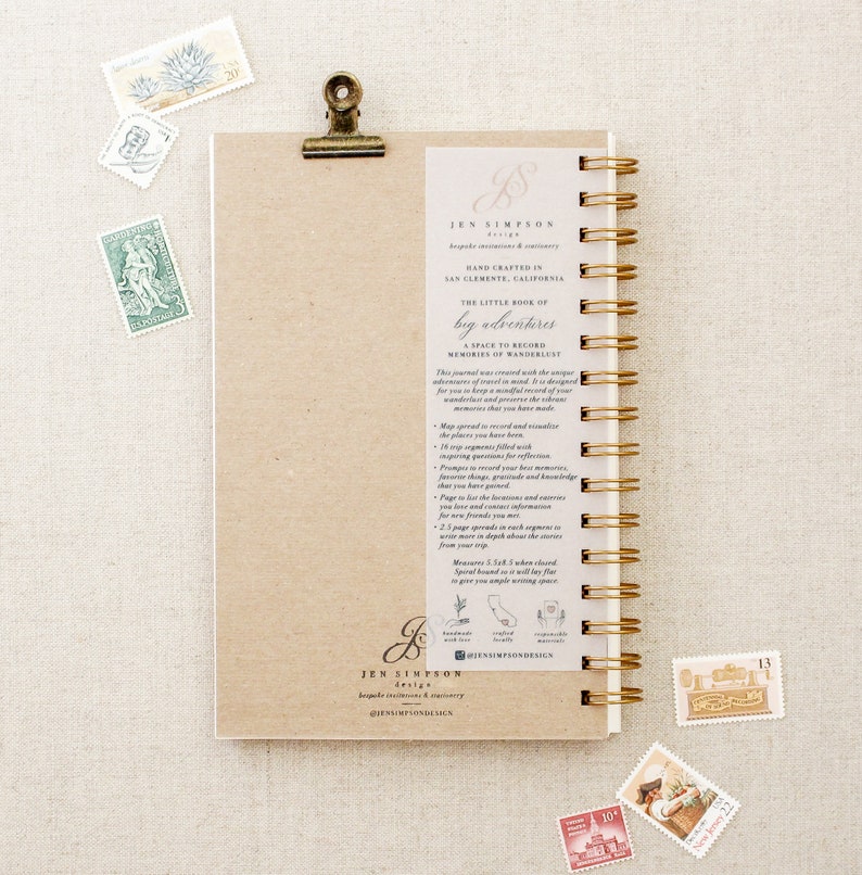 May include: A brown, spiral bound journal with a gold clip. The cover has a handwritten inscription that reads "The Little Book of Big Adventures: A Space to Record Memories of Wanderlust". The journal is designed to help you record your travel memories.