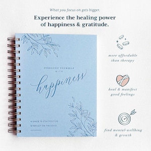 May include: A blue spiral bound journal with the words "Surround Yourself with Happiness" and a floral design. The journal is a space to stay positive and reflect on the good. The text "What you focus on gets bigger. Experience the healing power of happiness & gratitude." is at the top of the image.