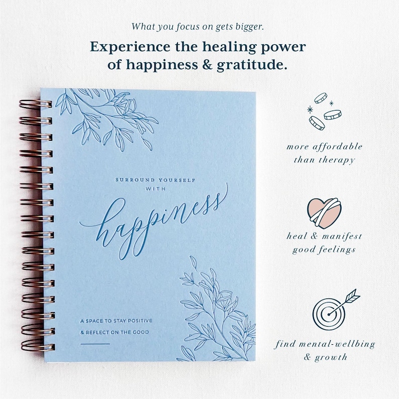 May include: A blue spiral bound journal with the words "Surround Yourself with Happiness" and a floral design. The journal is a space to stay positive and reflect on the good. The text "What you focus on gets bigger. Experience the healing power of happiness & gratitude." is at the top of the image.