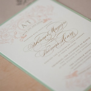May include: A wedding invitation with a light green border and a white background. The invitation features a decorative design with a floral motif and the text "AT" in a circle. The text "Together with their families" is written below the circle. The names "Anne Maguire" and "Thomas Henry" are written in cursive script below the text. The text "Request the pleasure of your company at their wedding" is written below the names.