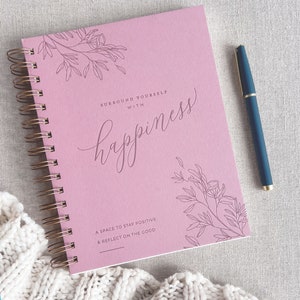 May include: A pink journal with a gold spiral binding. The cover features a floral design and the text "Surround yourself with happiness. A space to stay positive & reflect on the good."