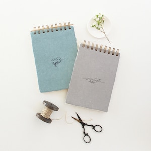 May include: Two spiral bound notebooks with a rustic paper texture. The notebook on the left is blue and says "notes" with a small floral design. The notebook on the right is gray and says "secrets" with a small floral design.
