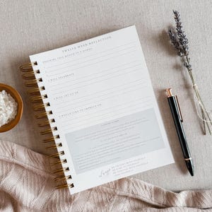 May include: A white spiral bound journal with gold rings. The cover has the text "Twelve Week Reflection" and includes prompts for self-reflection. The journal is lying on a beige surface with a pen and lavender flowers.
