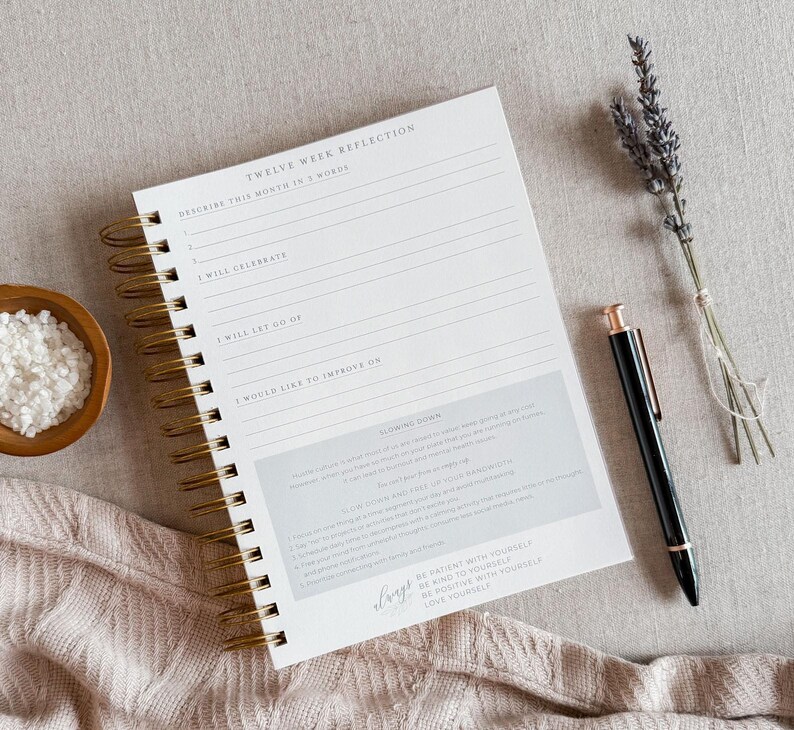 May include: A white spiral bound journal with gold rings. The cover has the text "Twelve Week Reflection" and includes prompts for self-reflection. The journal is lying on a beige surface with a pen and lavender flowers.