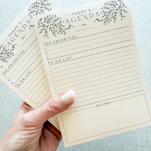 May include: Two cream-colored agenda notepads with black text and floral accents. The top of the notepad reads "TODAY'S AGENDA" with space for the date. Below are sections for "TOP PRIORITIES" and "TO DO LIST". The bottom has a section that reads "don't forget".