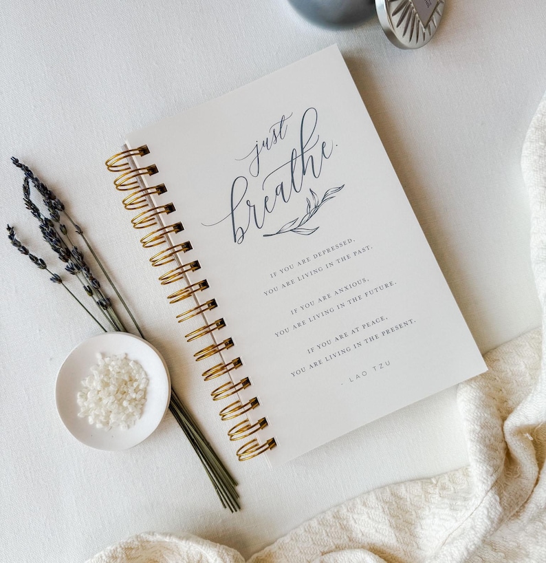 Pu&ograve; includere: Un taccuino a spirale con copertina bianca che riporta il testo Just breathe in un'elegante calligrafia, seguito da una citazione di Lao Tzu su depressione, ansia e pace. Il taccuino presenta una rilegatura a spirale dorata e una finitura opaca.