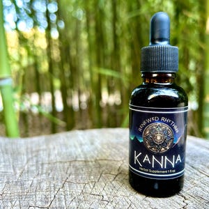 May include: A black glass bottle with a dropper, labeled "Renewed Rhythms Kanna Herbal Supplement 1 fl oz". The bottle is sitting on a wooden surface with a blurred background of green bamboo.