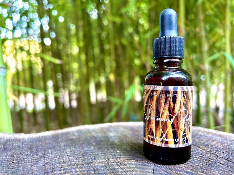 May include: A brown glass bottle with a dropper top and a label that reads "Shatavari Root" and "Renewed Rhythms". The bottle is sitting on a wooden surface with a blurred background of green bamboo.