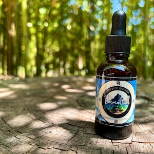 May include: A brown glass bottle with a black dropper and a label that reads "Brewed Rhythm Immortal." The label features a mountain scene within a circular design. The bottle sits on a weathered wooden surface, with a blurred green background.