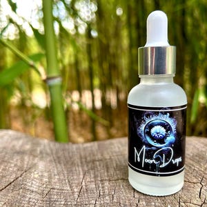 May include: A frosted glass bottle with a white dropper top and a silver collar. The bottle has a black label with the text "Moon Drops" in white script. The background is a blurred view of bamboo.