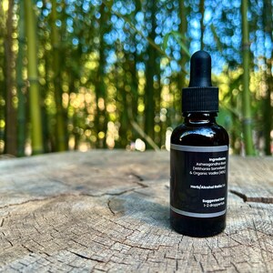 May include: A dark amber glass bottle with a black dropper top, set on a weathered wood surface. The label includes text about ingredients and suggested use. The background features blurred green bamboo stalks.