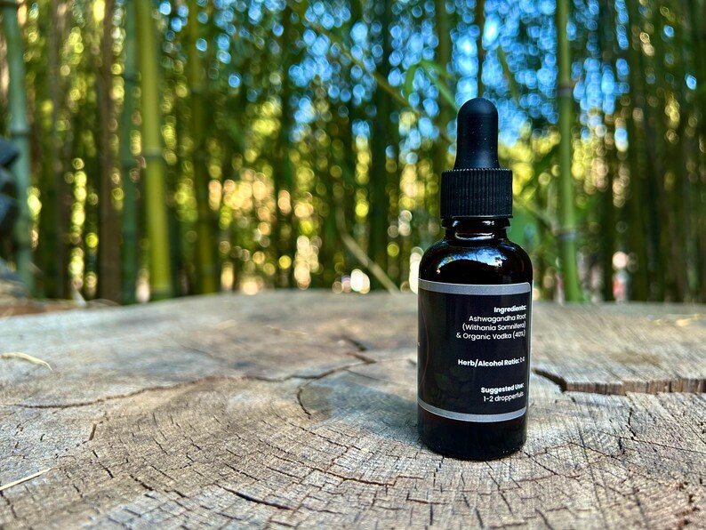 May include: A dark amber glass bottle with a black dropper top, set on a weathered wood surface. The label includes text about ingredients and suggested use. The background features blurred green bamboo stalks.