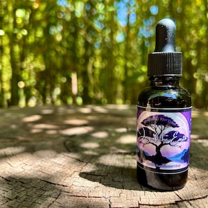 May include: A dark glass bottle with a black dropper, featuring a purple label with white text and a tree graphic. The bottle is set on a weathered wooden surface with a blurred green background.