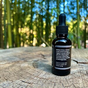 May include: A dark brown glass bottle with a black dropper, resting on a weathered wooden surface. The bottle has a label with white text that reads: "Reduce stress, balance mood, boost energy..." The background is blurred green bamboo.