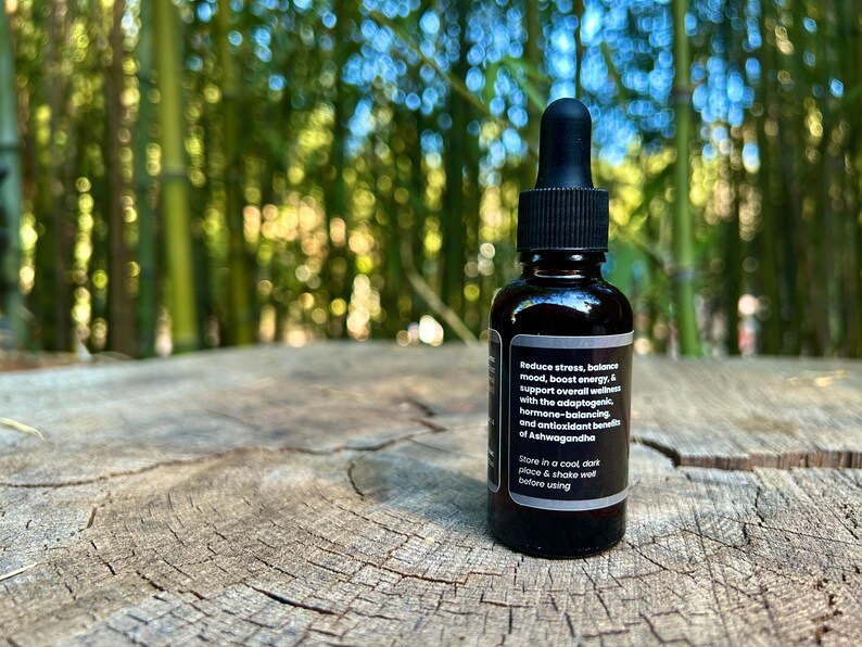 May include: A dark brown glass bottle with a black dropper, resting on a weathered wooden surface. The bottle has a label with white text that reads: "Reduce stress, balance mood, boost energy..." The background is blurred green bamboo.