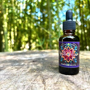 May include: A small, dark brown glass bottle with a dropper top. The bottle is labeled "Wild Muse" and features a colorful mandala design. The bottle is sitting on a wooden surface.