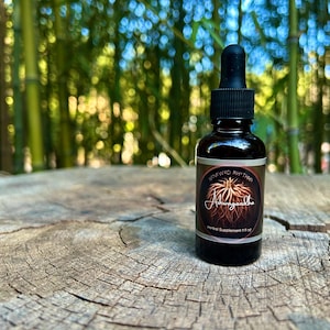 May include: A dark brown glass bottle with a black dropper cap, labeled "Renewed Rhythm Ashwagandha Herbal Supplement 1 fl oz." The bottle sits on a weathered, textured wooden surface, with a blurred green bamboo forest in the background.
