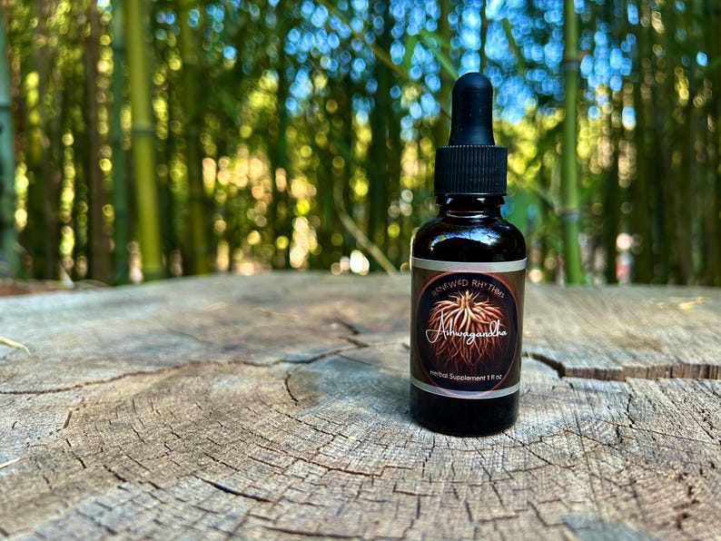 May include: A dark brown glass bottle with a black dropper cap, labeled "Renewed Rhythm Ashwagandha Herbal Supplement 1 fl oz." The bottle sits on a weathered, textured wooden surface, with a blurred green bamboo forest in the background.