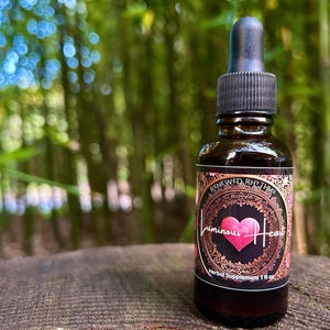 May include: A dark brown glass bottle with a black dropper top and a gold label that says "Renewed Rhythms Luminous Heart Herbal Supplement 1 fl oz". The label has a gold ornate design and a red heart in the center.
