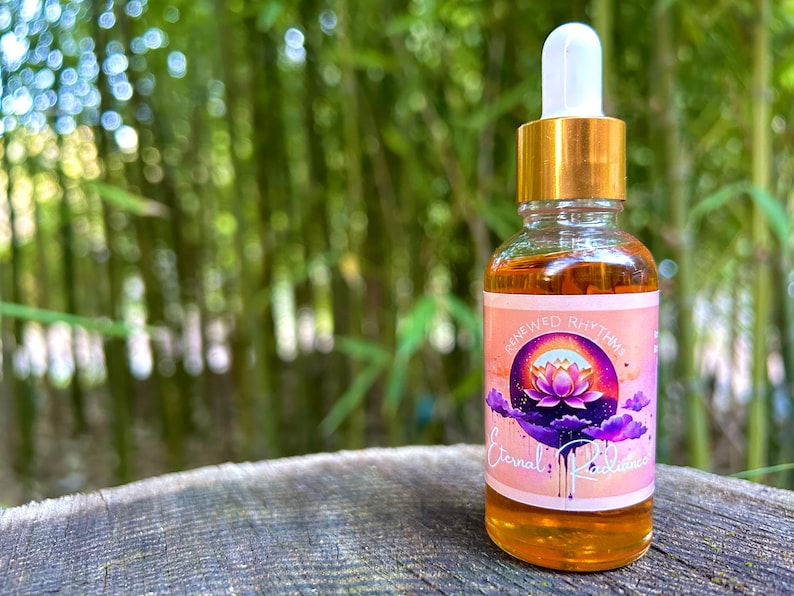 1oz Eternal Radiance beauty serum bottle with a pink & purple label. A vibrant lotus flower sits at the center, surrounded by soft purple clouds, symbolizing renewal and radiance. The design is calming, with a focus on natural beauty & glowing skin.
