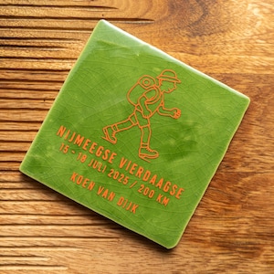 Op de afbeelding: Groene keramische tegel met een witte omtrek van een wandelaar met een rugzak. De tegel heeft de tekst "Nijmeegse Vierdaagse 15-18 juli 2025 / 200 km Koen van Dijk" in oranje.