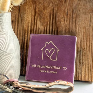 Może przedstawiać: Kwadratowa, fioletowa płytka ceramiczna ze złotym tekstem i konturem domu z sercem w środku. Tekst brzmi "WILHELMINASTRAAT 23" i "Feline & Jeroen". Widoczny jest również biały wazon z suszonymi kwiatami i pęk kluczy.