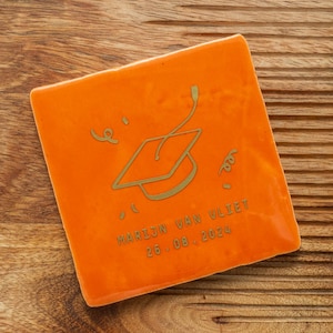 Op de afbeelding: Oranje vierkante tegel met een gouden illustratie van een afstudeerhoed en de tekst "MARIJN VAN VLIET 26.08.2024".