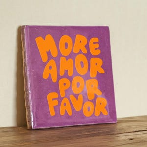 Può includere: Piastrella quadrata viola con il testo "MORE AMOR POR FAVOR" in lettere arancioni a forma di bolla.