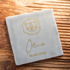 Może przedstawiać: Biała płytka ceramiczna z złotą ilustracją lwa i imieniem "Otis" napisanym kursywą. Data "10.03.2024" jest napisana pod imieniem.