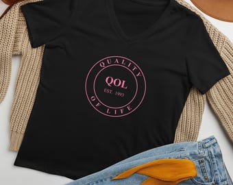 May include: Black V-neck t-shirt with a pink circular graphic that reads "QUALITY OF LIFE" with "QOL EST. 1993" in the center. The shirt is displayed with blue jeans, a tan sweater, and a brown purse.