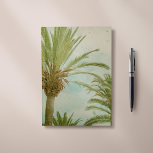 Peut inclure: Un carnet avec une illustration de palmier et un stylo. Le carnet présente une image de style vintage de palmiers sur fond de ciel bleu clair. Un stylo noir et argenté est placé à droite du carnet.