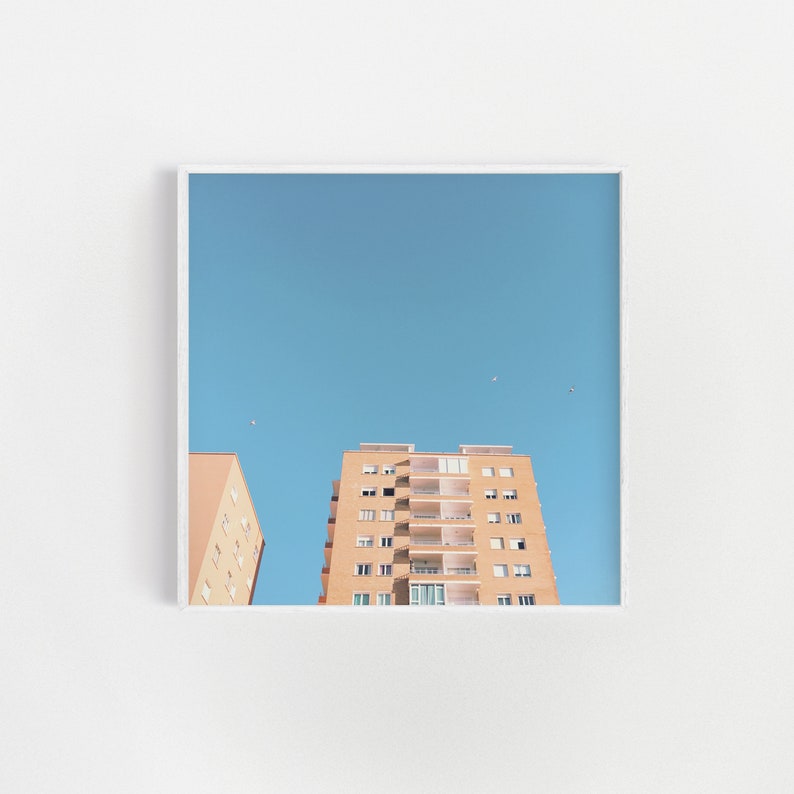 May include: A photograph of a tall, light brown apartment building with a blue sky in the background. The building has many windows and balconies. There are a few birds flying in the sky.